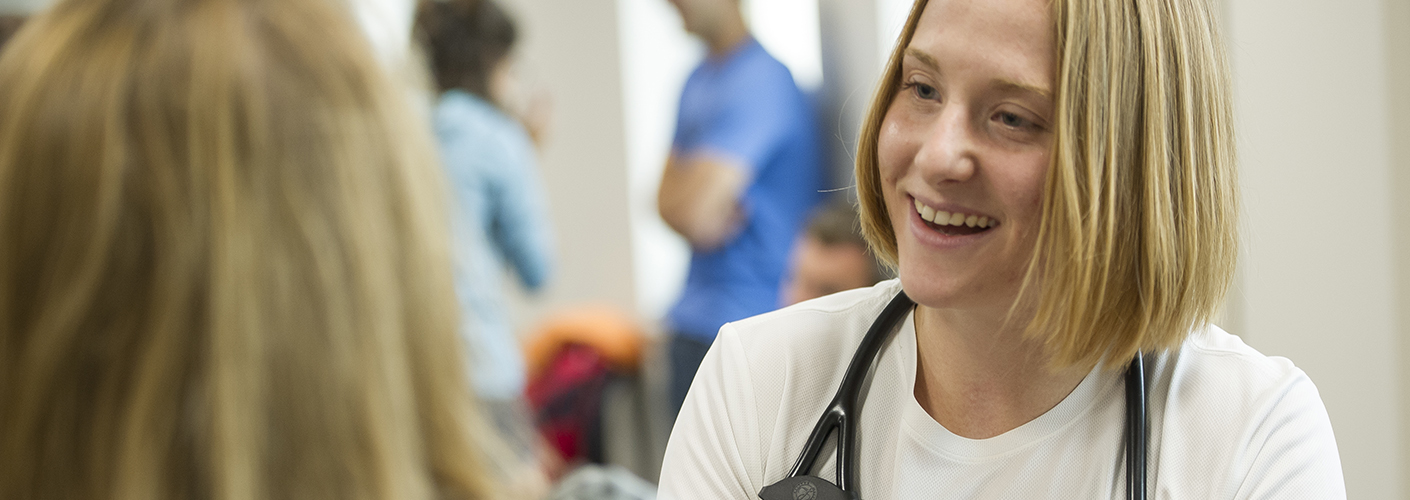 A young woman with a stethoscope around her neck chats with a patient.