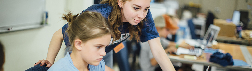 A young woman guides a student through a task on a computer.