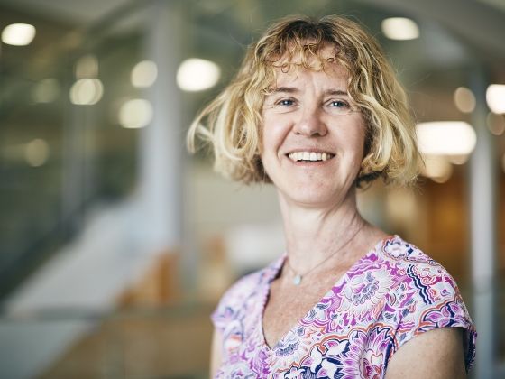 Smiling woman with short, wavy blonde hair wearing a short-sleeve top with a purple floral pattern.