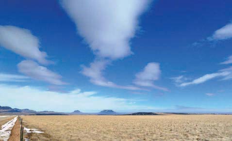 Elongated clouds stretch across a bright blue sky above a flat, open grassland.  The smooth, layered cloud formations appear lens-shaped and aligned with atmospheric airflow over the landscape.