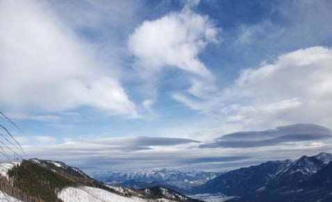 Several smooth, lens-shaped dark gray clouds stretch horizontally across the lower altitudes of a blue sky. Above them, fluffy white clouds dot the sky. Snow-covered peaks and forested slopes frame a broad valley in the distance. 