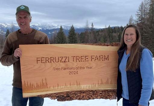 a photograph of a man and woman standing outside in the snow with trees and mountains in the background. They are holding a large wooden sign that reads, Ferruzzi Tree Farm Tree Farmers of the Year 2024. 