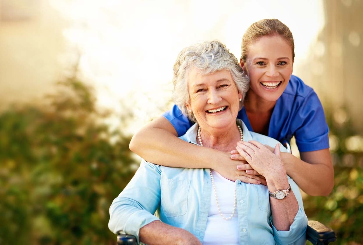 A smiling older female in a light blue button-down shirt over a white shirt sitting with a smiling younger female wearing a medium blue shortsleeved shirt bent hugging the older female around the neck from behind.