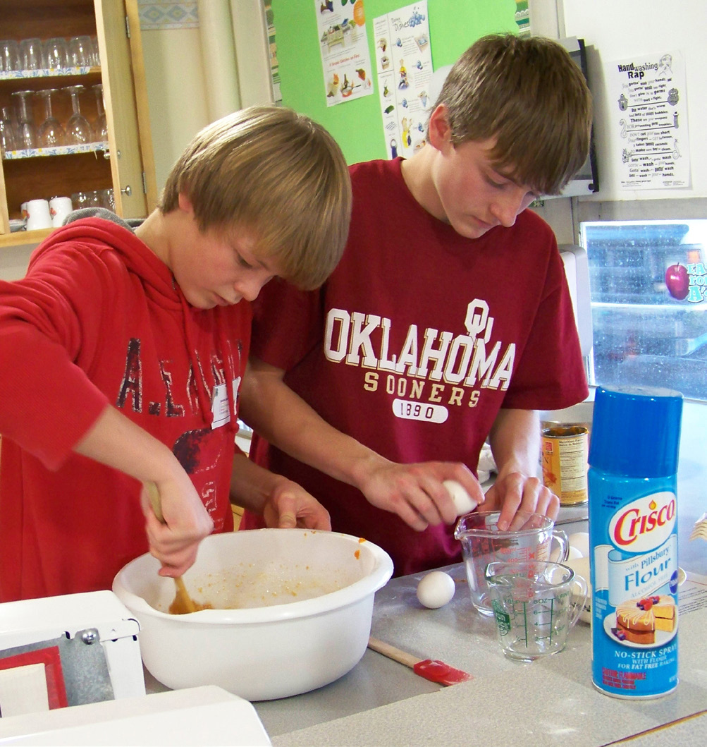 A boy cracks eggs into a bowl while the other mixes.