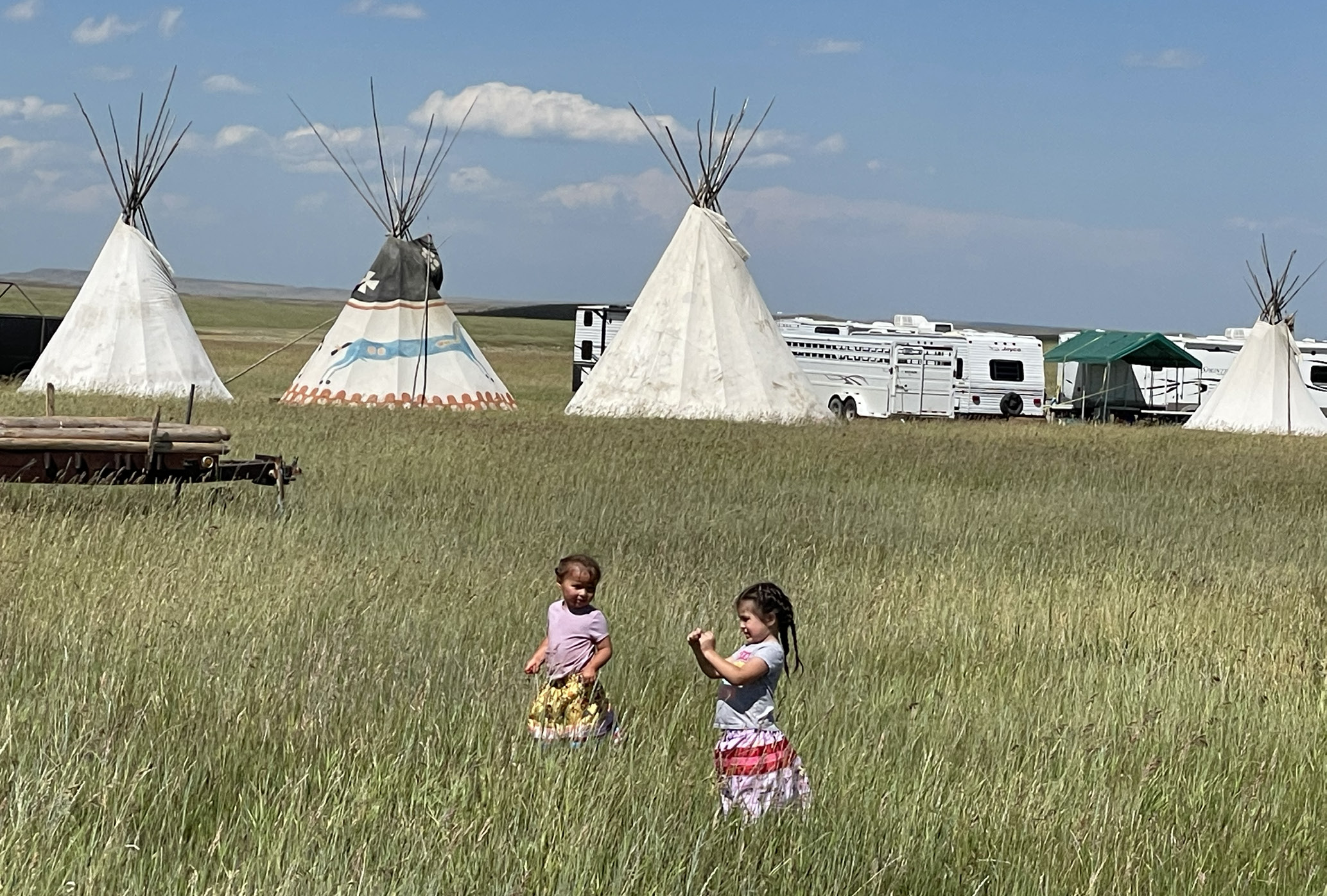 Two children play in a field of tall green grass. Tipi's stand in the background.