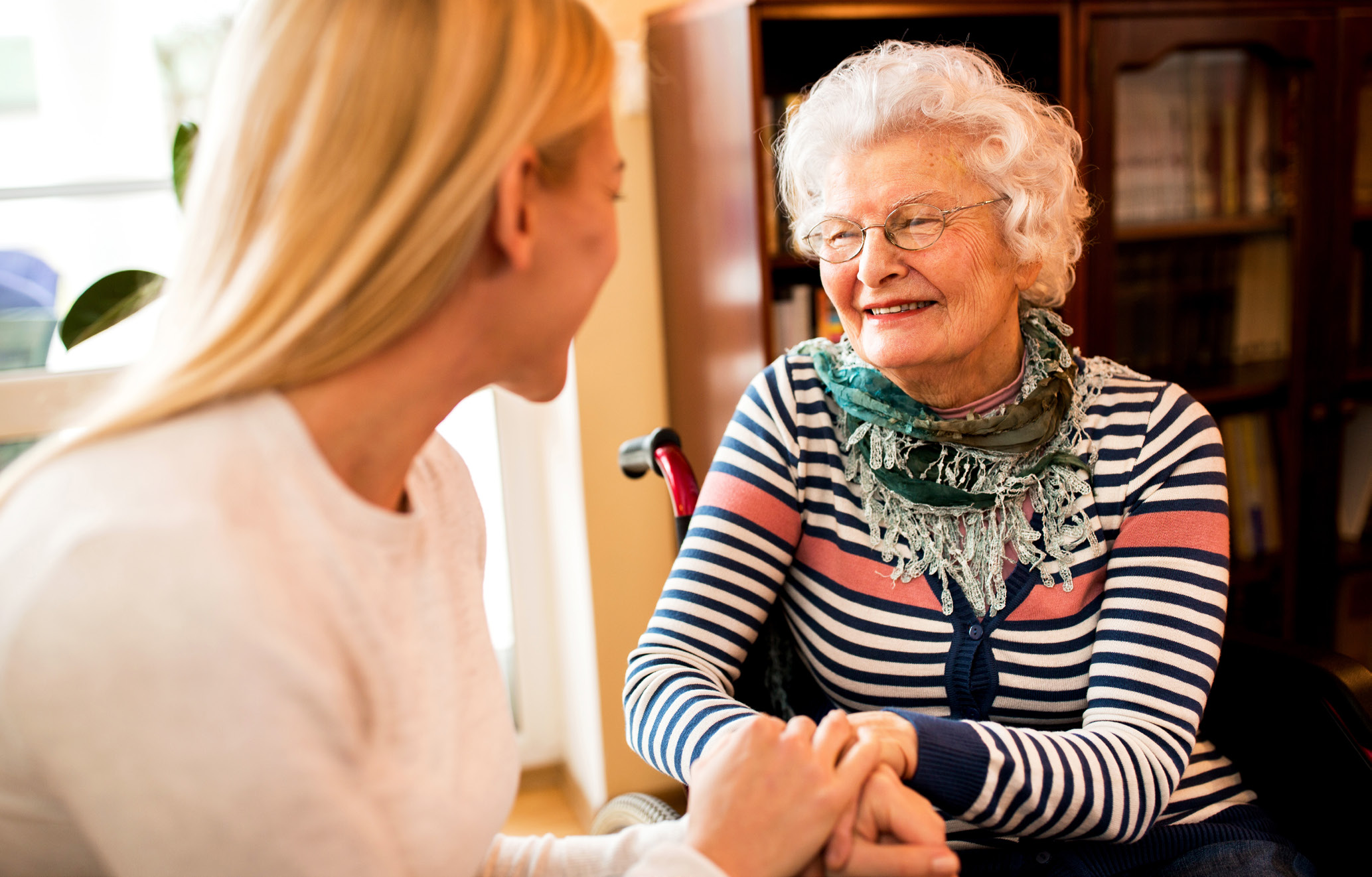 Two women sit holding hands, one is aged with white hair and a wheel chair and the other is younger.