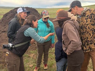 Participants gathered to examine a soil sample while standing near compost piles in open grassland.
