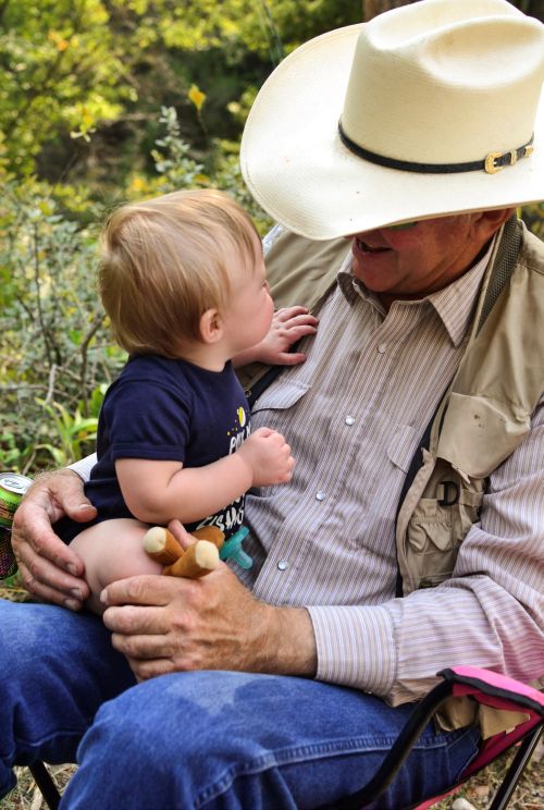 An older man in a cowboy hat and fishing vest holds a small child on his lap. They are looking at each other and smiling