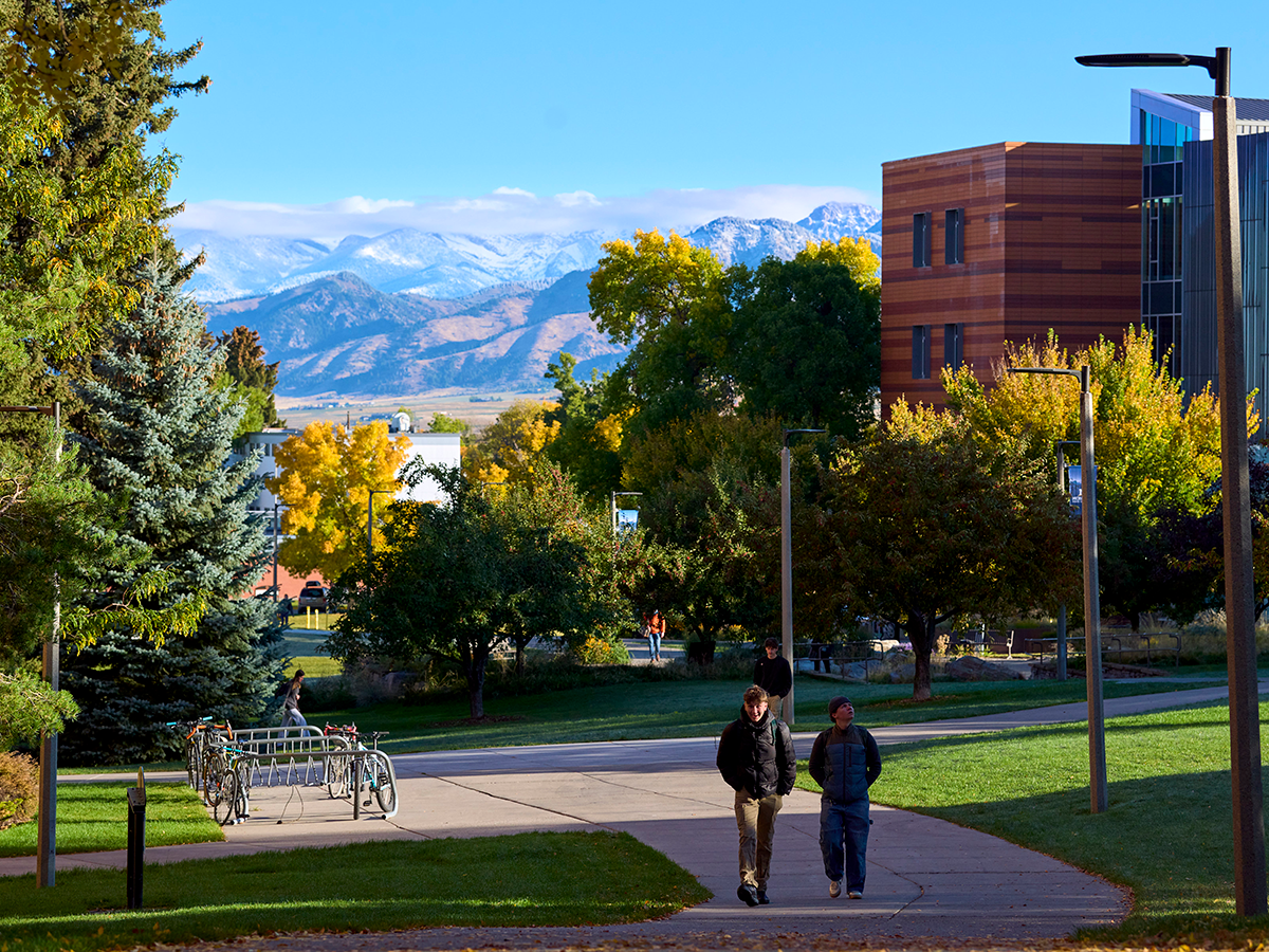 Two students walking on a paved area with brick buildings and trees in the background. There are snowy mountans further in the background.