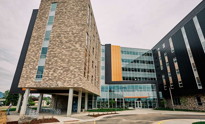 Metal, glass, concrete and brown brick make up the exterior of Hyalite Hall. The six-story residence hall is designed in the shape of a capital letter “H”. This photo shows the top of the “H” shape which is the main entrance of the residence hall. There is covered bike parking on the ground floor of the left wing of the building.