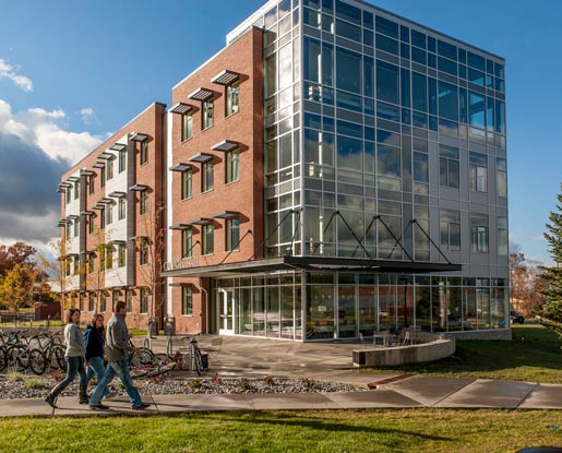 Three people walk past Gallatin Hall, a four-story rectangular building made of glass, brick and metal. One side of the building is constructed entirely from glass.
