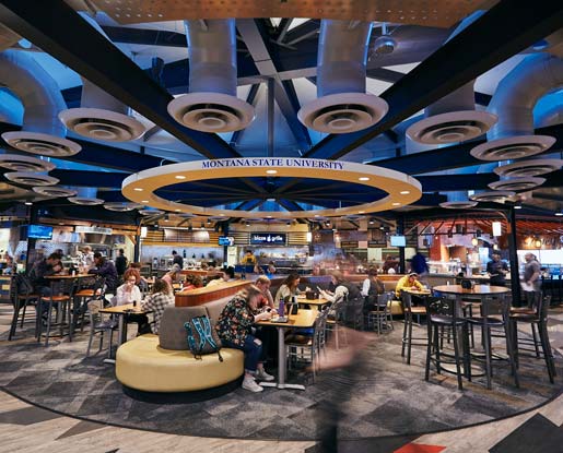 A contemporary, colored photo shows a bustling dining area at Montana State University's Miller Dining Hall. Students are shown walking quickly by as they appear slightly blurry. Several students are shown at various tables. Some students are eating with friends while others are working on laptops, looking at phones, and listening to music. A key feature of this building's construction and highlighted in this image is the futuristic ventilation system that makes a circular pattern on the ceiling above the main dining area.