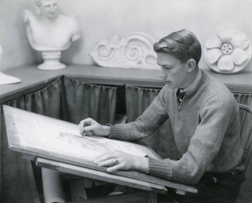 A black-and-white photo shows a student sketching a scale model statue using a pencil. He is sitting at a drafting desk and wearing a quarter-zip sweater, collared shirt and tie. There are three additional scale model statues in the background of the image. They are displayed on a table up against the wall.