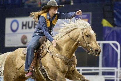 Rodeo rider in western attire competes on a galloping palomino horse inside an arena.