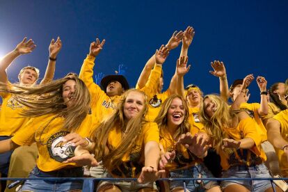 University fans in yellow shirts enthusiastically cheer and reach forward during an outdoor evening sports event.
