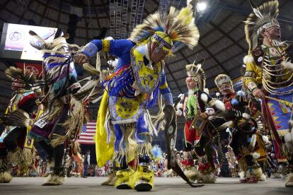 Dancers in colorful regalia perform a traditional Native American dance at an indoor powwow event.