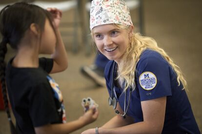 Nursing student in scrubs with Montana State University patch talks with a child holding a sticker during community outreach.