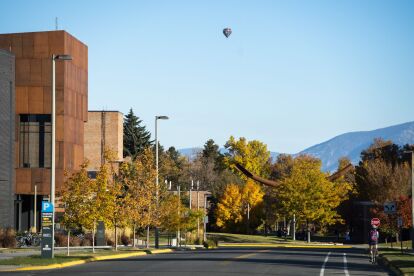 Campus street lined with autumn trees and buildings, a cyclist rides near a stop sign under a clear sky with a hot air balloon.