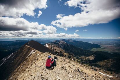 Person in outdoor gear sits on rocky ridge overlooking expansive mountain range under partly cloudy sky.
