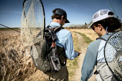 Two anglers walk along a dirt path carrying fishing nets and gear on a clear day.