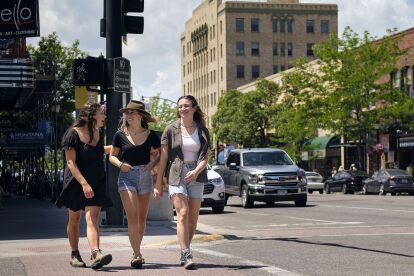 Three young people walk across a city street on a sunny day with a tall building and trees in the background.