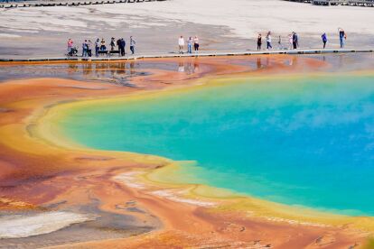 Visitors walk along a boardwalk beside a vibrant geothermal pool with blue, green, yellow, and orange hues.