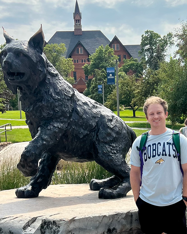 A person wearing a gray BOBCATS crewneck sweatshirt, standing next to a bobcat statue