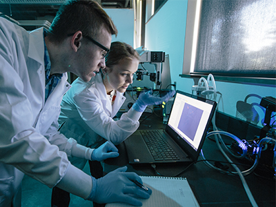 Two students in lab coats look at a computer in a lab