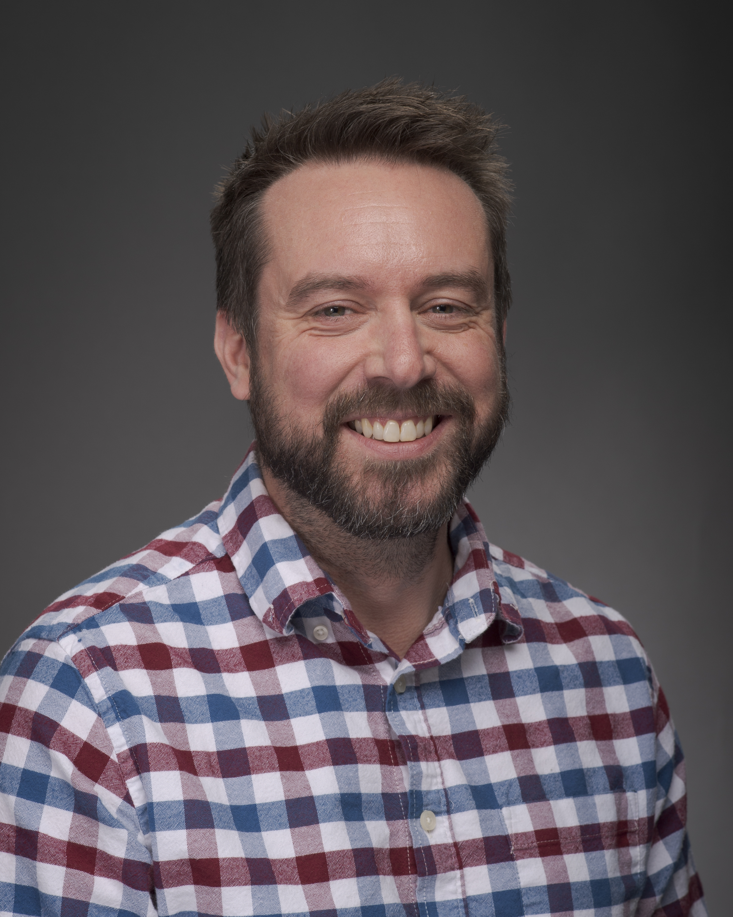 Portrait of a person with short dark hair and facial hair, wearing a plaid button up shirt