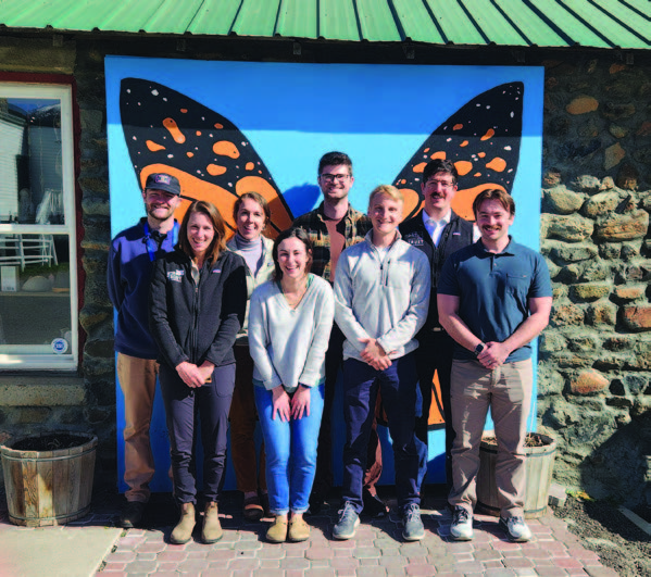 A grou of eight women and mean standing in front of a large butterfly mural, smiling and looking straight ahead.
