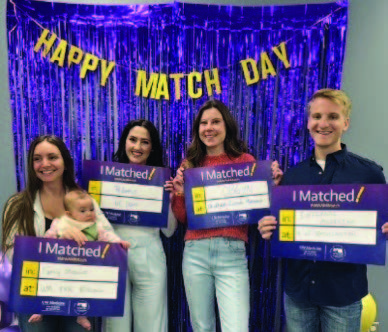 A group of three women and one man smiling and holding signs that say "I Matched." A gold banner over their heads reads "Happy Match Day" against a purple background. One of the women is holding a baby.