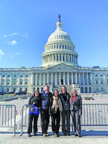 A group of five women and one man standing in fron tof the U.S. Capitol, smiling and looking straight ahead. 