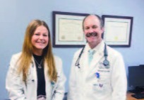 A female medical student standing next to a male doctor, both wearin white lab coats and smiling straight ahead.