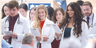 A group of smiling medical students wearing their white cotas and stethoscopes looking down and reading from a piece of paper.